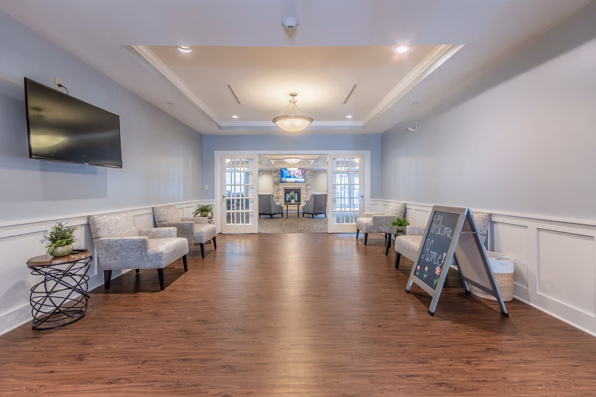 Bright senior living facility seating area with chairs along the walls, a wall-mounted TV, and a small 'Welcome Home' sign near the entrance.