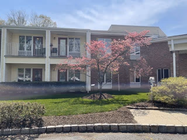 Exterior view of a two-story assisted living facility building with balconies and windows. In front of the building, there is a landscaped area with green grass, a blooming pink tree, and some shrubs under a partly cloudy blue sky.