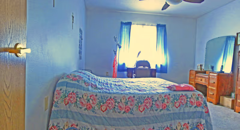 A bedroom with a bed covered in a floral quilt, a wooden dresser with a large mirror, a chair in front of a window with blue curtains, and a ceiling fan above.