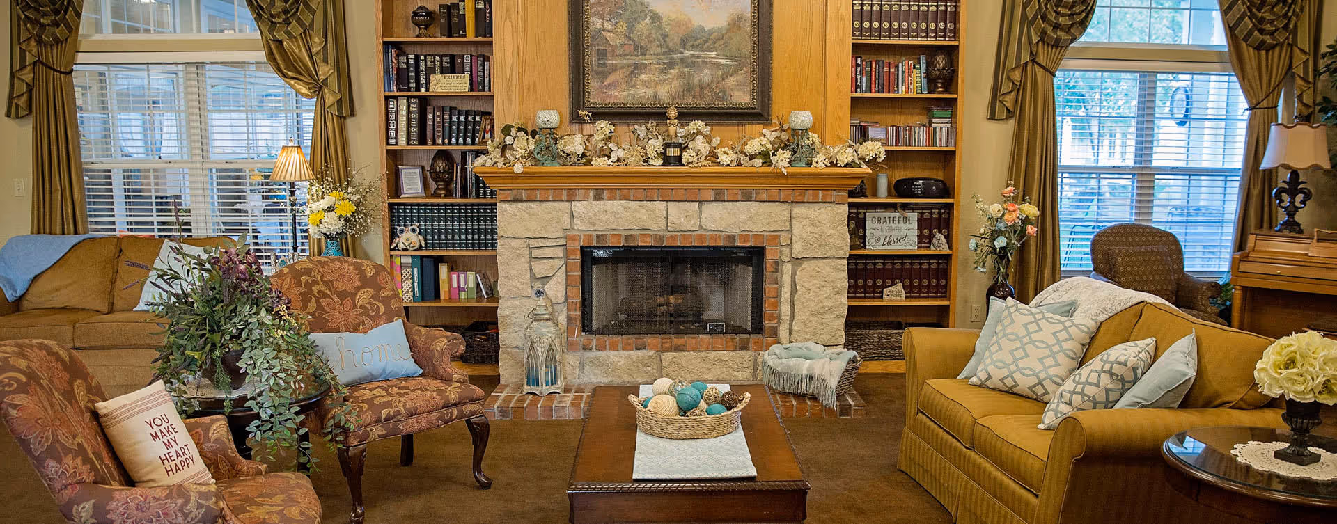A cozy living room with a stone fireplace in the center, flanked by built-in wooden bookshelves filled with books and decorative items. There are two large windows with green curtains on either side of the fireplace. The room features comfortable seating including a mustard yellow sofa with patterned pillows, two floral armchairs, and a brown sofa. A wooden coffee table with a decorative basket sits in the middle of the room. Various plants and flowers add a touch of greenery and warmth.