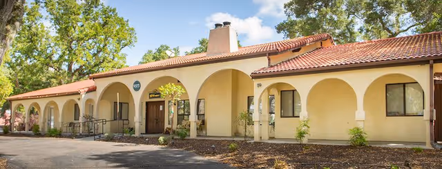 Exterior view of Ingleside Assisted Living Inc building featuring a beige stucco facade with multiple arches, a red tile roof, and a chimney. The building is surrounded by trees and has a paved area in front.