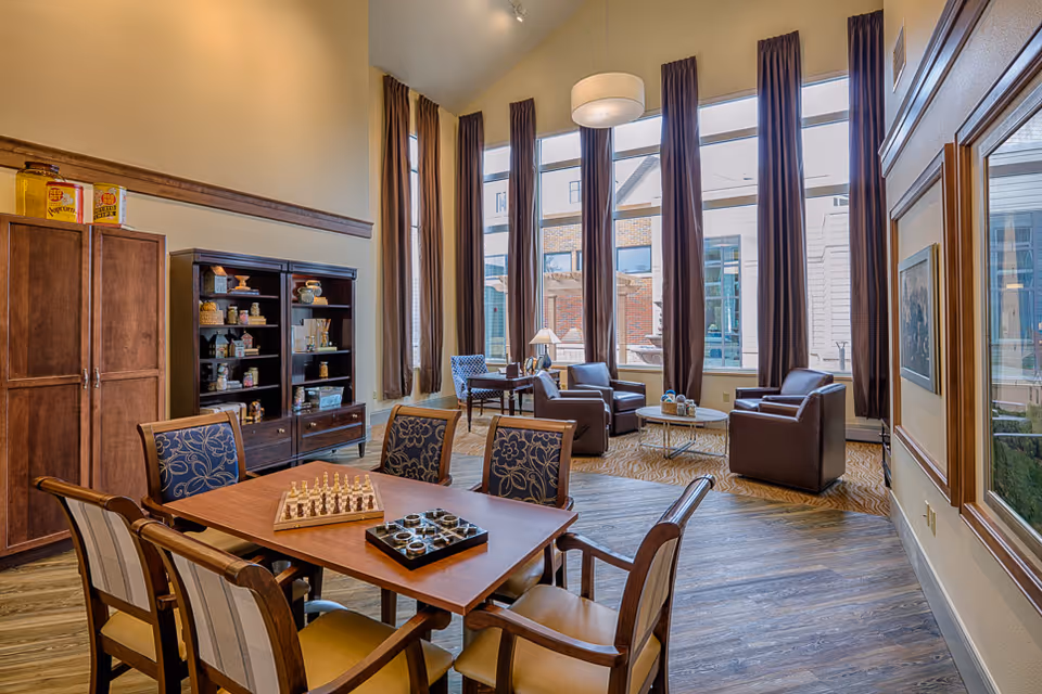 A bright and spacious common area in a senior living facility with tall windows covered by long curtains, several armchairs arranged around a coffee table, a wooden table with chairs featuring a chessboard and a checkers game, and shelves with decorative items and games.