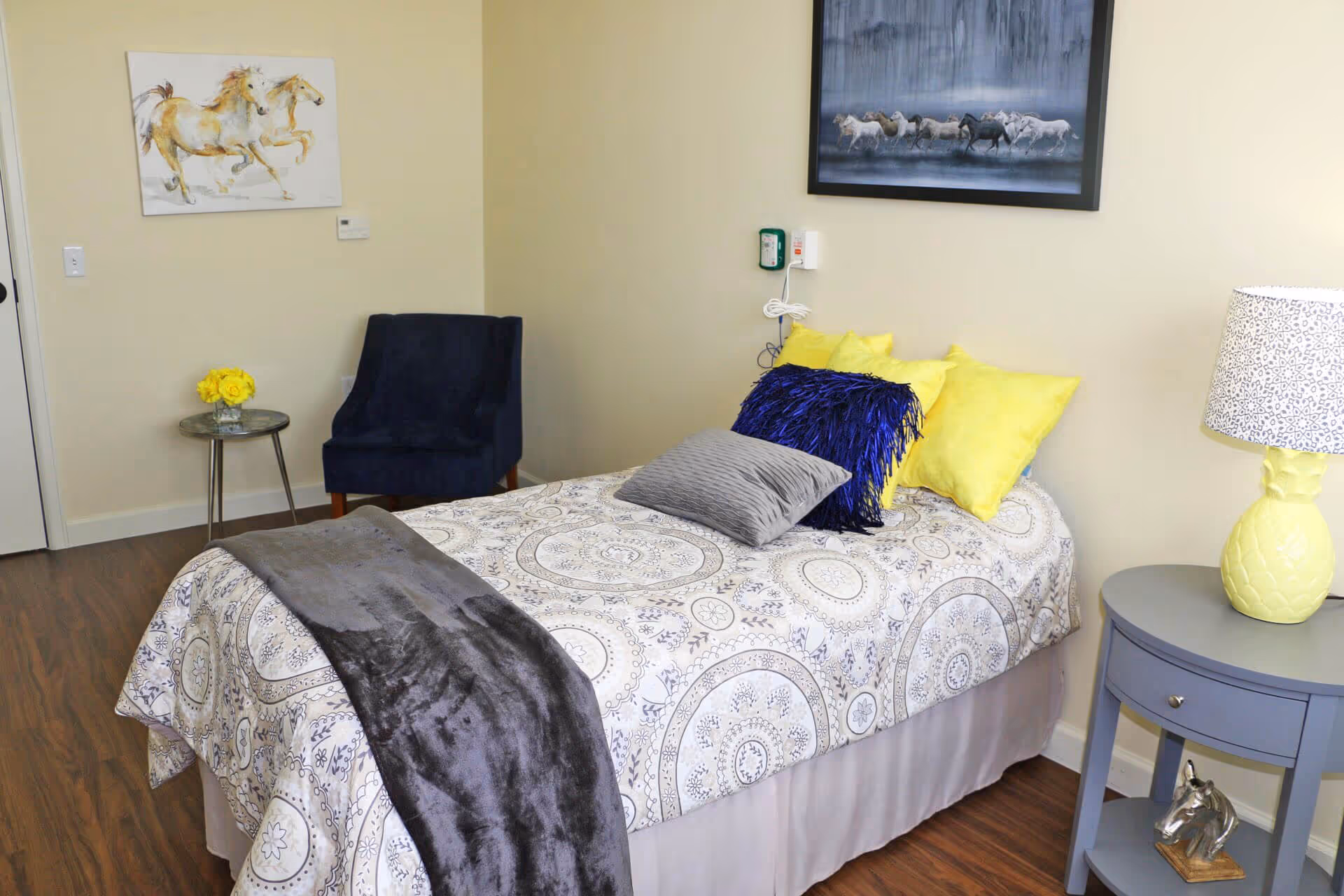 A cozy bedroom with a single bed covered in a patterned bedspread and several colorful pillows including yellow and blue. A gray throw blanket is draped over the bed. There is a small round side table with a yellow lamp and a decorative horse head figurine. A dark blue armchair sits next to a small glass table with a vase of yellow flowers. Two paintings of horses hang on the light-colored walls. The floor is wooden.
