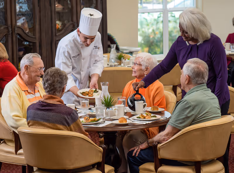A group of elderly people sitting around a dining table in a retirement living facility, being served food by a chef in a white uniform and a woman pouring coffee into a cup. The setting is warm and inviting with large windows in the background.