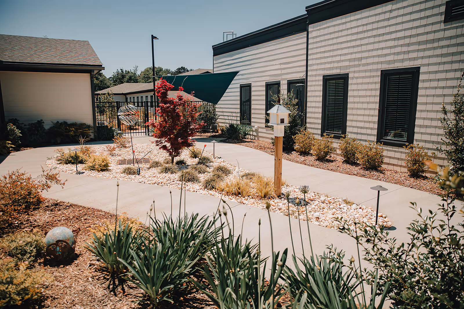 Outdoor garden area at Aiken Memory Care featuring a paved walkway surrounding a landscaped section with various plants, a small red tree, decorative garden ornaments, and a birdhouse mounted on a wooden post. The building exterior with multiple windows is visible on the right side under a clear blue sky.