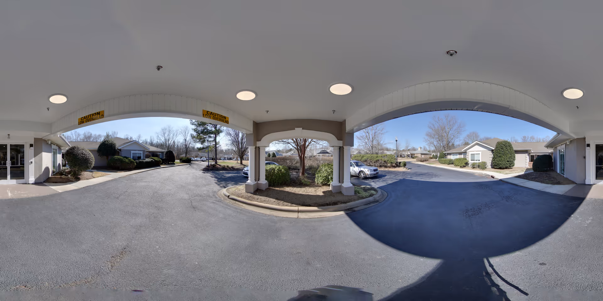 Covered driveway entrance at Mint Hill Senior Living facility with a circular driveway, landscaped bushes, and parked cars under a white ceiling with round recessed lights and caution wet floor signs.