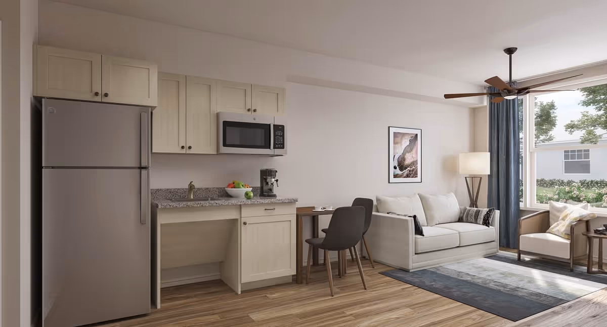 Open-plan studio living area with a small kitchenette on the left, a sofa and chairs by a large window on the right.