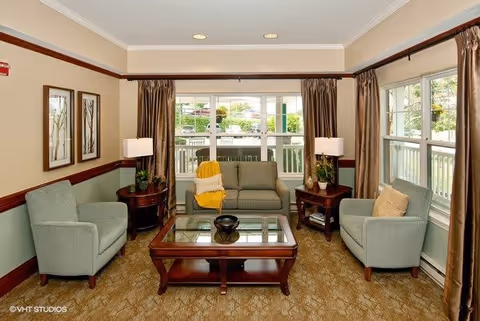 A cozy living room with a beige sofa and two light green armchairs arranged around a wooden coffee table with a glass top. The room has beige walls with a green lower half and dark wood trim. Two side tables with lamps and plants flank the sofa. Large windows with brown curtains let in natural light and offer a view of an outdoor patio area.