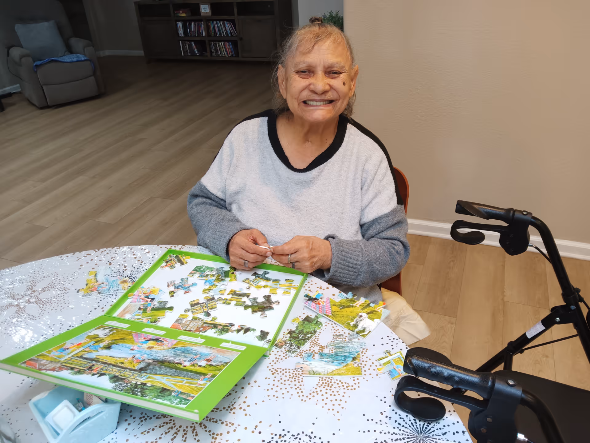An elderly woman smiling while sitting at a table working on a jigsaw puzzle. The table has a white tablecloth with a decorative pattern. A walker is positioned next to her. In the background, there is a recliner chair and a bookshelf with DVDs or books.