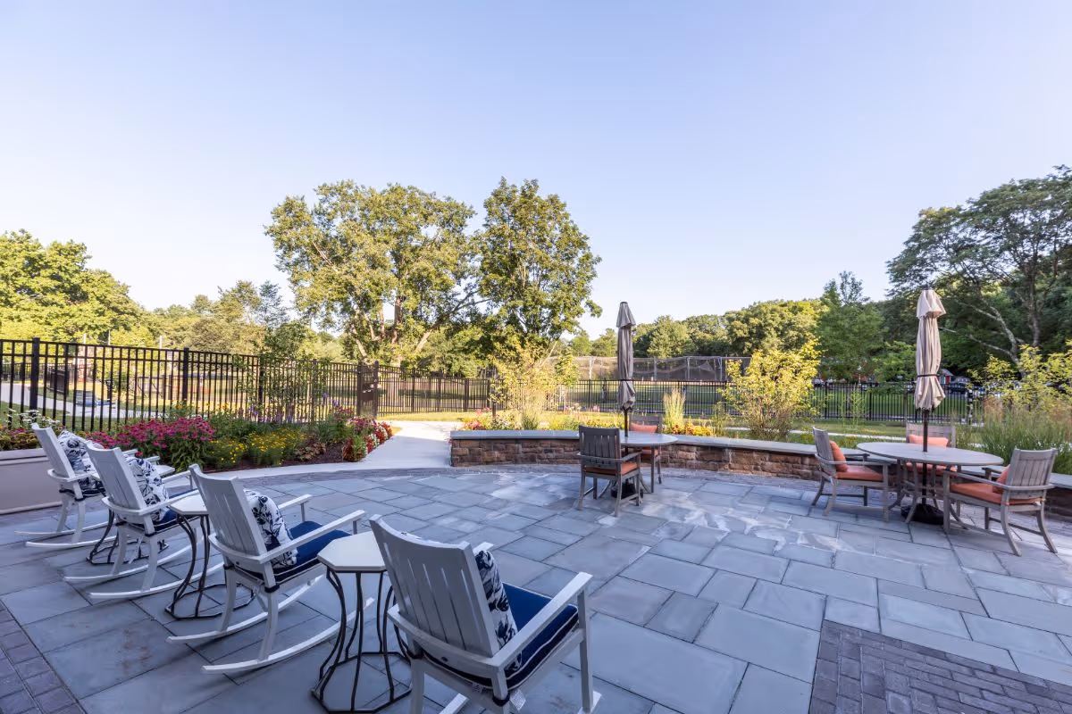 Outdoor patio area with white rocking chairs and small tables on a stone tiled floor. There are round tables with chairs and closed umbrellas. The patio is surrounded by a low stone wall and flower beds, with a black metal fence and trees in the background under a clear blue sky.