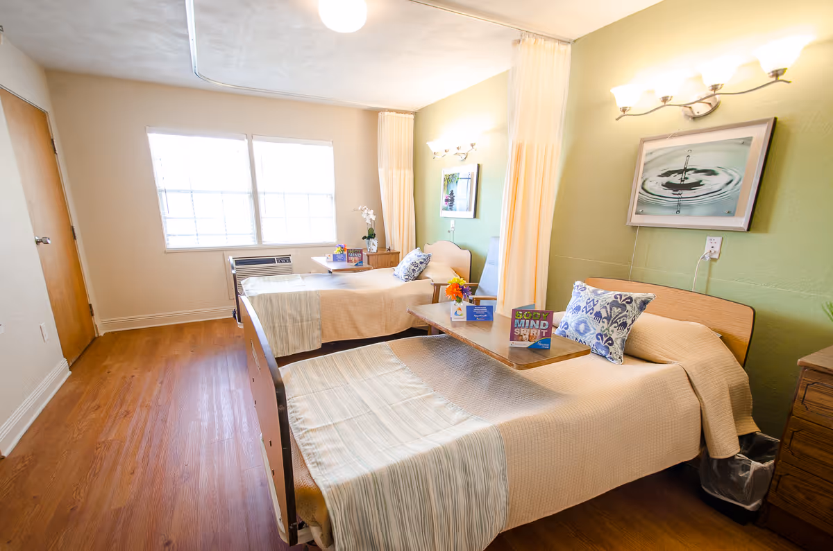 Two neatly made twin beds in a bright senior care bedroom with bedside tables, privacy curtains, and wall art.