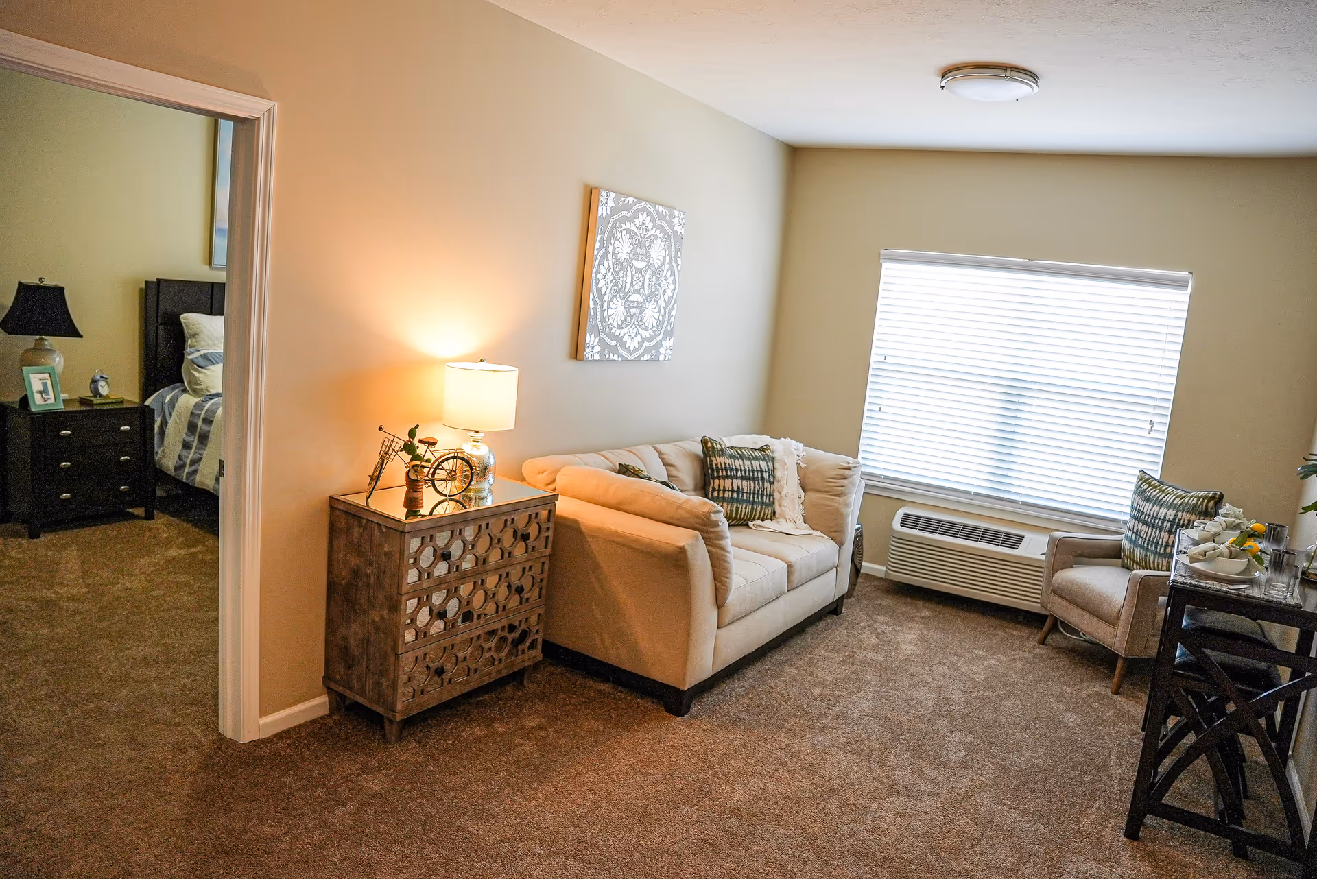 Cozy living room with a beige loveseat, armchair, decorative side table with a lamp, and a window with blinds.