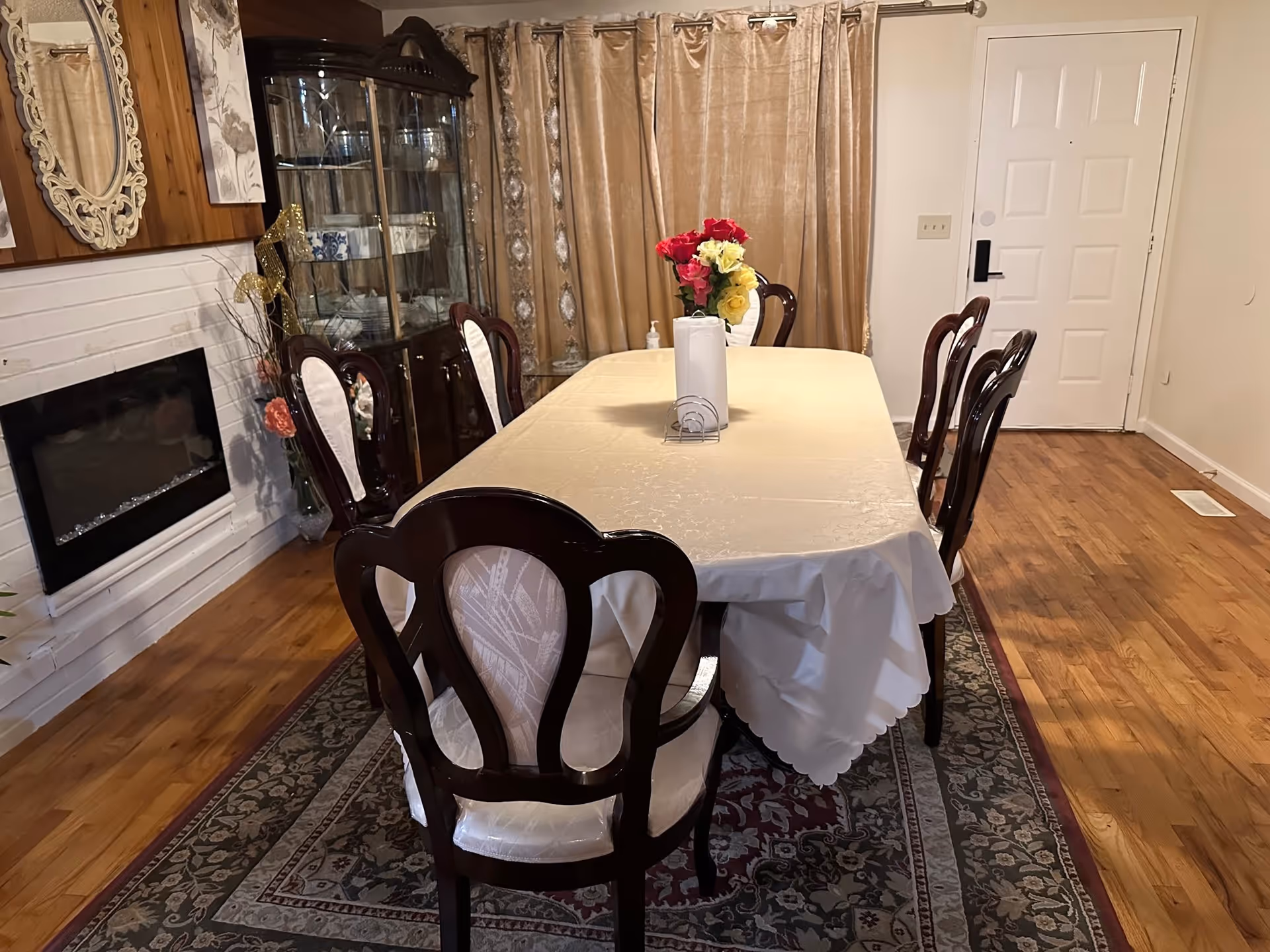 A dining room with a rectangular table covered with a cream-colored tablecloth, surrounded by six wooden chairs with white cushioned backs and seats. On the table, there is a vase with red, pink, and yellow flowers and a paper towel holder. The room has hardwood floors with a patterned area rug under the table. In the background, there is a glass-front china cabinet filled with dishes, beige curtains, a white door, and a wall-mounted electric fireplace with a decorative mirror above it.