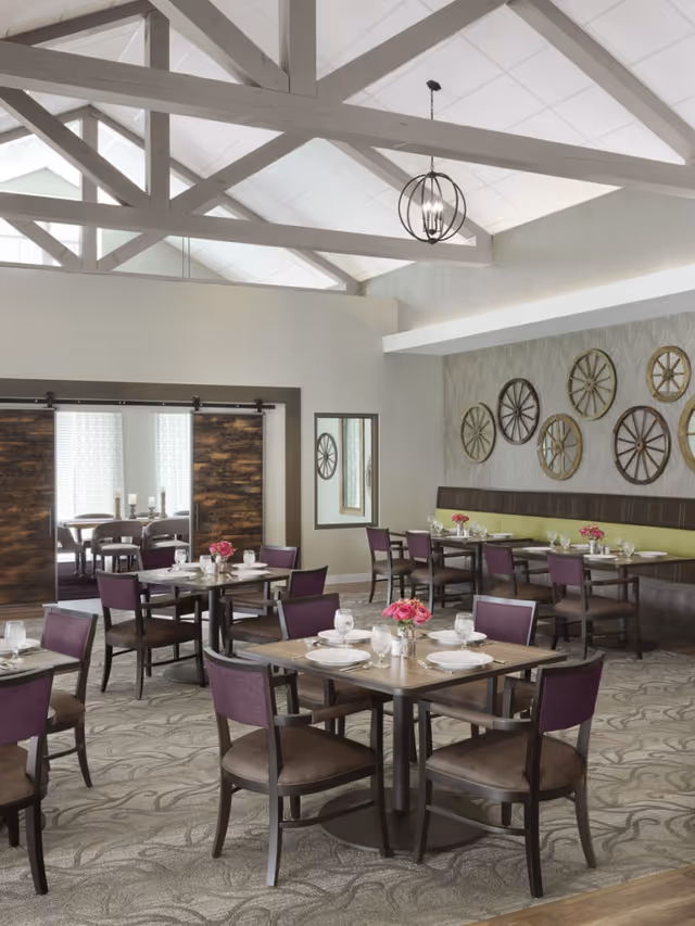 A bright and spacious dining room with several tables set for meals. Each table has four chairs with purple backs and brown seats, white plates, glasses, and pink flower centerpieces. The room features a high ceiling with exposed white beams and a modern hanging light fixture. One wall is decorated with various wooden wagon wheel designs, and there is a large mirror and sliding barn-style doors in the background.