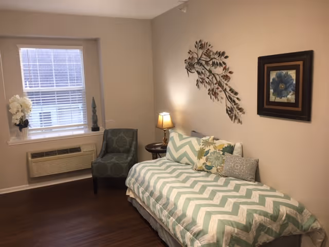 A cozy room with a single bed covered in a chevron-patterned bedspread in shades of green and white. The bed has several decorative pillows. Next to the bed is a small round wooden side table with a lamp on it. A green upholstered chair is positioned near a window with blinds. The walls are beige and decorated with a metal wall art piece shaped like branches with leaves and a framed floral painting. The floor is dark wood.