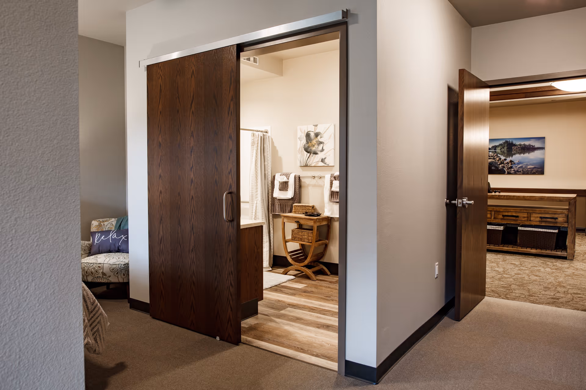 View of an interior hallway in a senior living facility showing a partially open dark wooden sliding door leading to a bathroom with towels and a small wooden table. To the right, an open door reveals a room with a wooden table, baskets underneath, and a landscape picture on the wall. A cushioned chair with a pillow that says 'relax' is partially visible on the left.