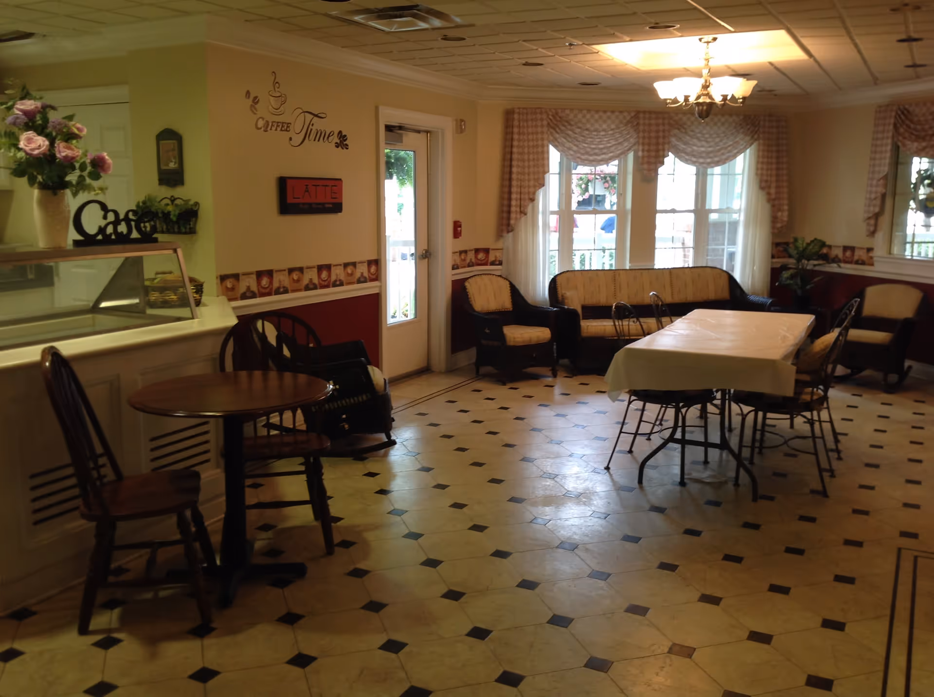Interior view of a cozy cafe area in Greenbriar Healthcare Center with a small round wooden table and chairs on the left, a counter with a glass display case, floral decorations, and a wall decal that says 'Coffee Time'. In the background, there is a rectangular table covered with a white tablecloth surrounded by chairs, cushioned wicker seating near large windows with checkered curtains, and a chandelier hanging from the ceiling.