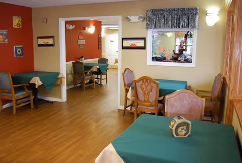 Interior view of a dining area with several tables covered with green tablecloths and wooden chairs. The room has light-colored walls with some framed artwork and a window with a floral curtain. The floor is wooden, and there is a small decorative clock on one of the tables.
