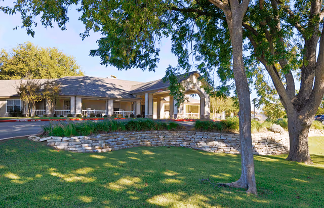 Exterior view of Sodalis Temple facility showing a single-story building with a covered entrance, surrounded by green grass, trees, and landscaping with stone retaining walls under a clear blue sky.
