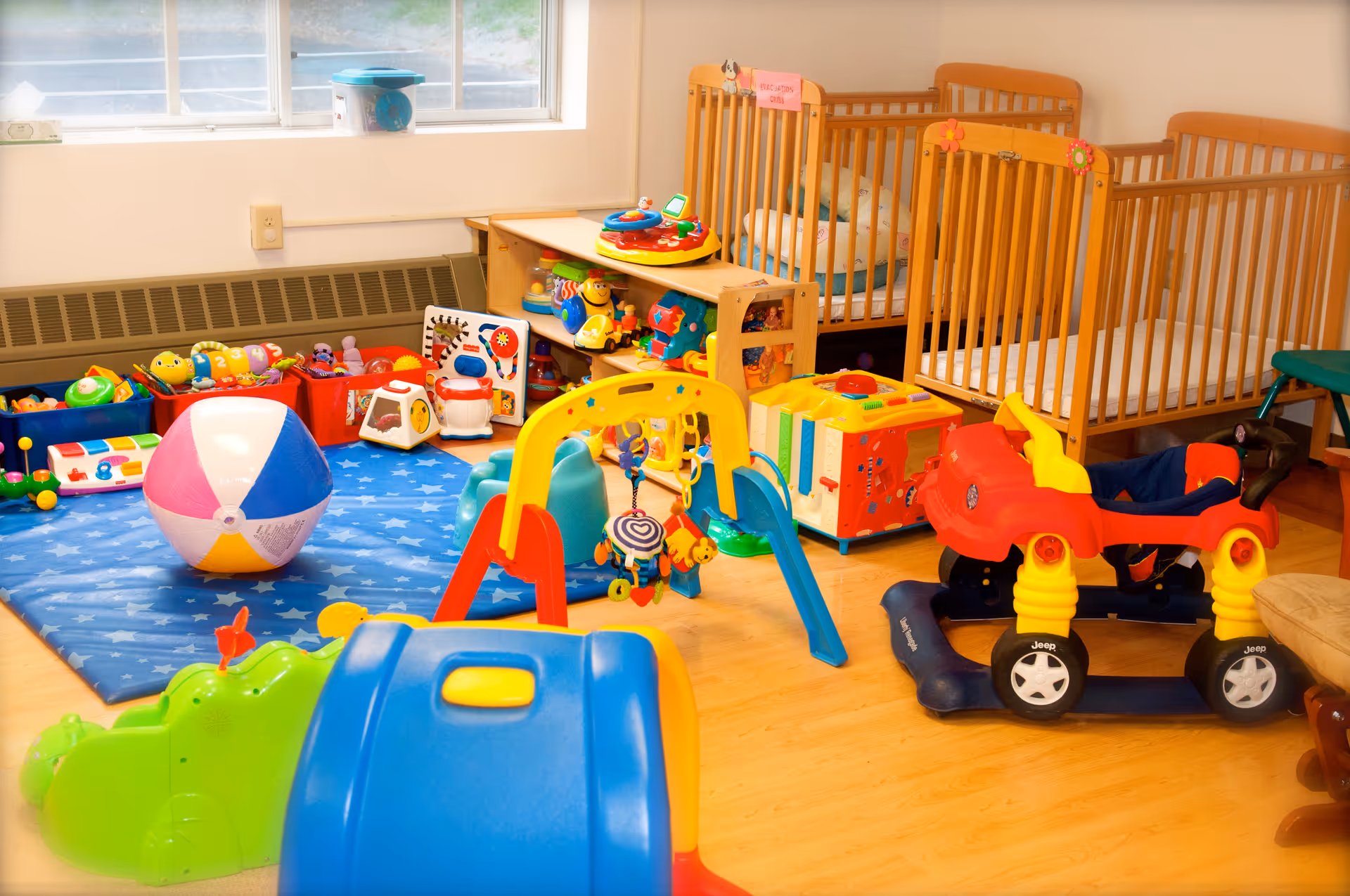 A brightly lit playroom with various colorful children's toys including a beach ball, toy car, play gym, and shelves filled with toys. There are also three wooden cribs lined up against the wall under a window.