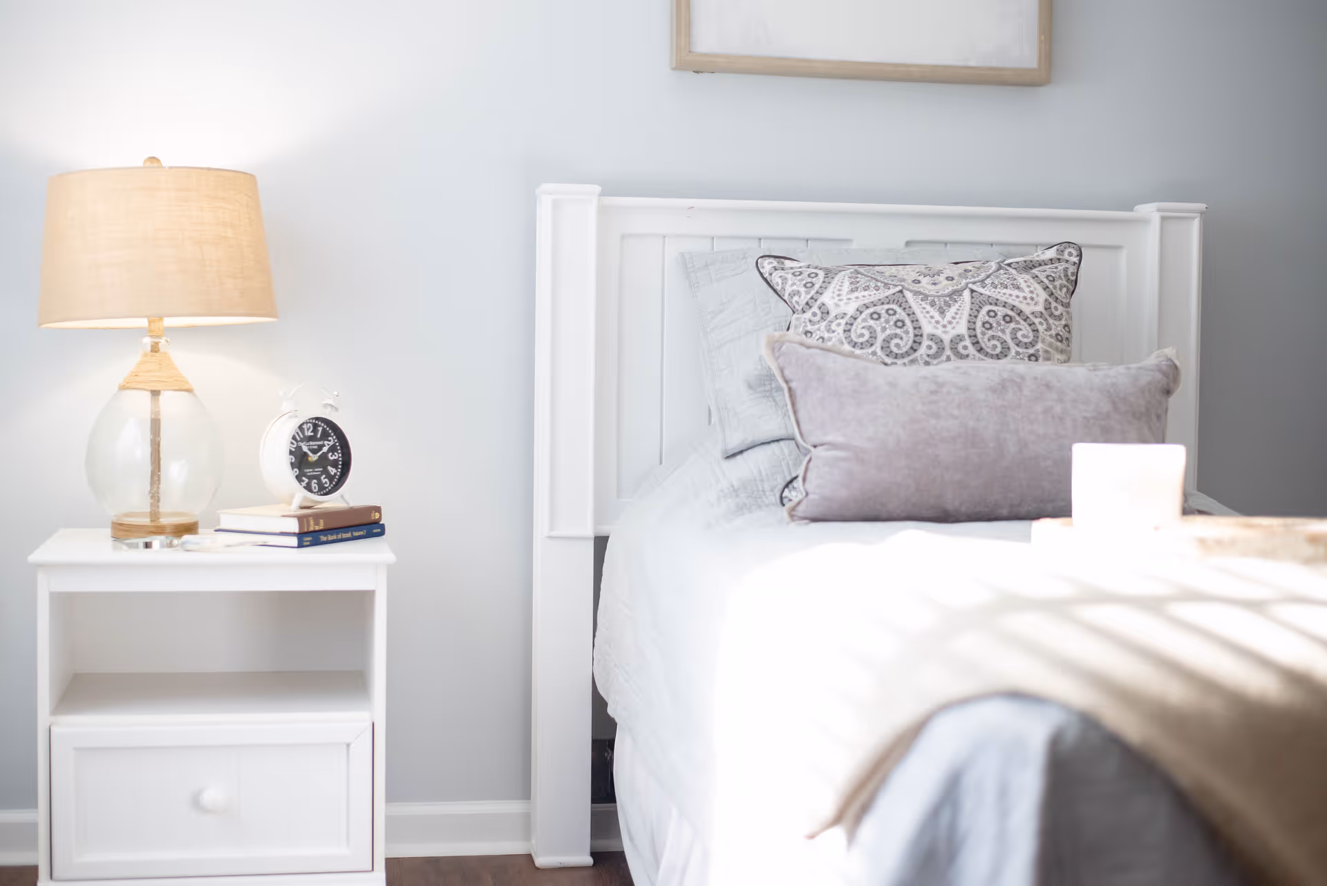 A bright bedroom scene with a white bed, patterned pillows, and a nightstand holding a lamp and alarm clock.