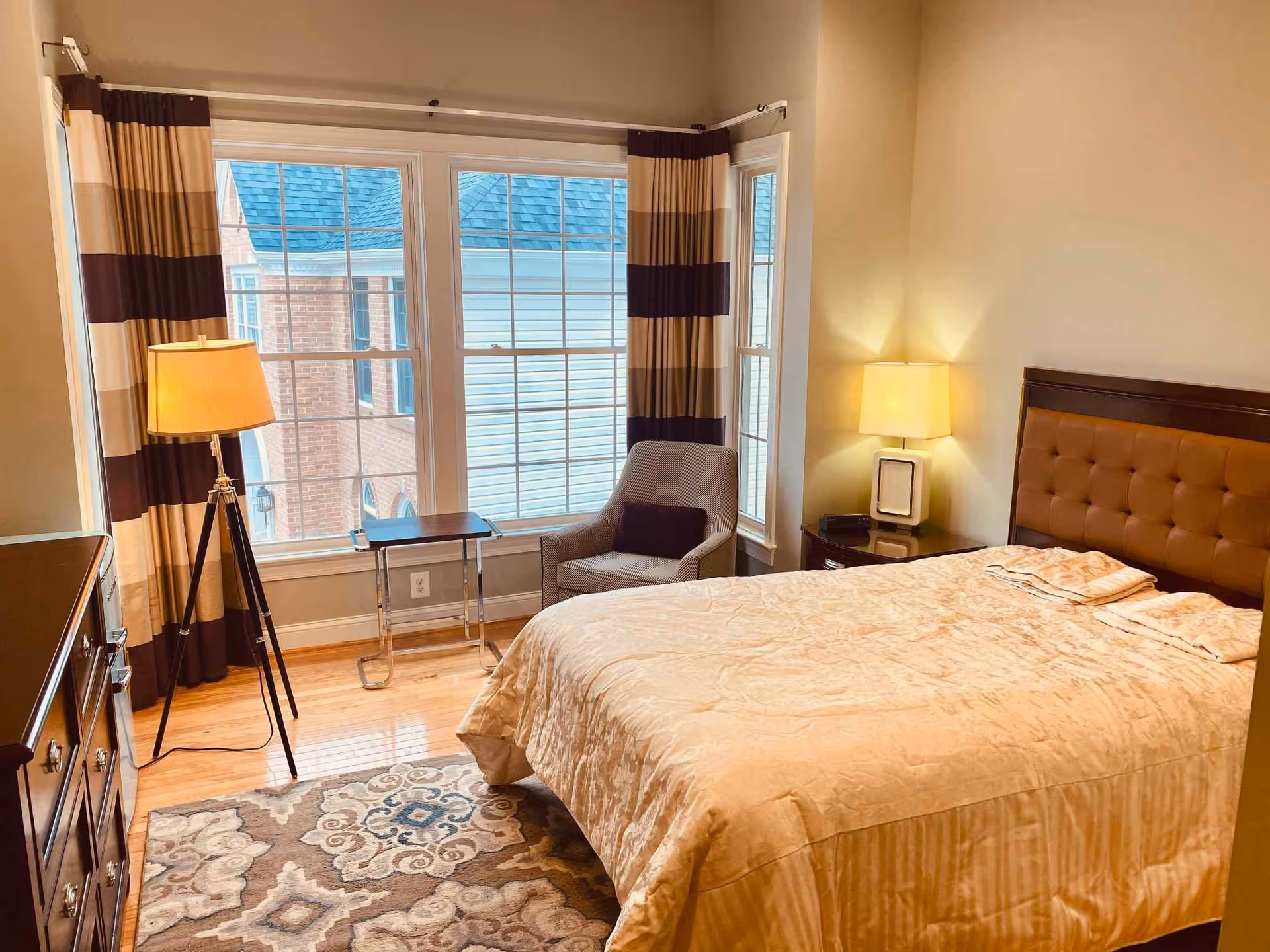 Sunlit bedroom with a bed, upholstered headboard, armchair by large windows with striped curtains, dresser, lamps, and a patterned rug.
