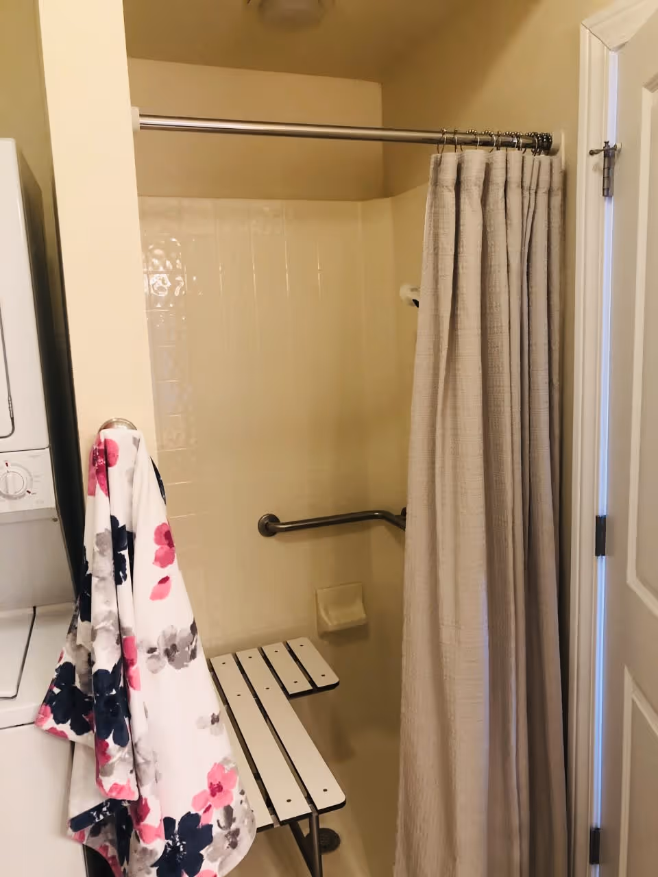 A bathroom shower area with a beige shower curtain partially closed. Inside the shower, there is a white foldable shower bench and a metal grab bar attached to the tiled wall. To the left of the shower, a floral-patterned towel hangs on the side of a stacked washer and dryer unit. The bathroom walls are painted a light beige color.