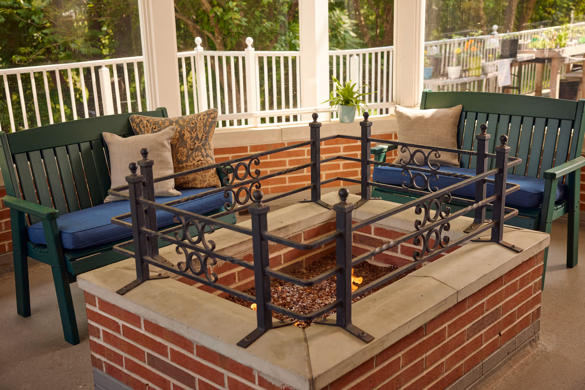 A cozy outdoor seating area with two green benches featuring blue cushions and decorative pillows, positioned around a brick fire pit with a black wrought iron safety railing. The area is enclosed with white railings and surrounded by greenery.