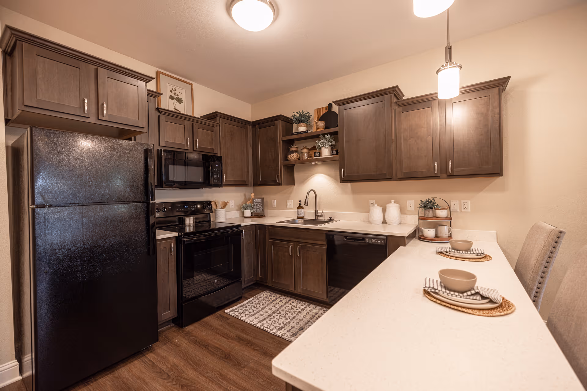 A modern kitchen with dark wood cabinets, black refrigerator, stove, microwave, and dishwasher. The kitchen features a white countertop with two place settings, a sink, and decorative plants and jars on the shelves and counter. The floor is wooden, and the lighting includes ceiling and pendant lights.
