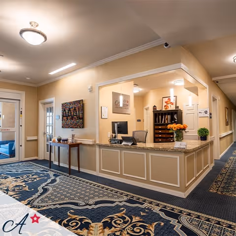 Reception area of All American Assisted Living At Enfield featuring a front desk with a computer, a vase of orange flowers, and a decorative cabinet behind the desk. The area has beige walls, patterned carpet, and framed artwork on the walls. There is a hallway leading further into the facility and a small table with decorative items near the entrance.