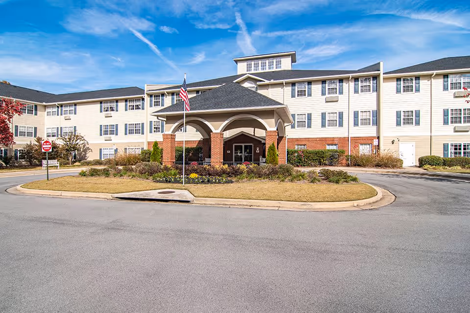 Exterior view of Creekside Pines Retirement Community building with a covered entrance supported by brick pillars, an American flag on a flagpole, landscaped garden beds, and a clear blue sky.