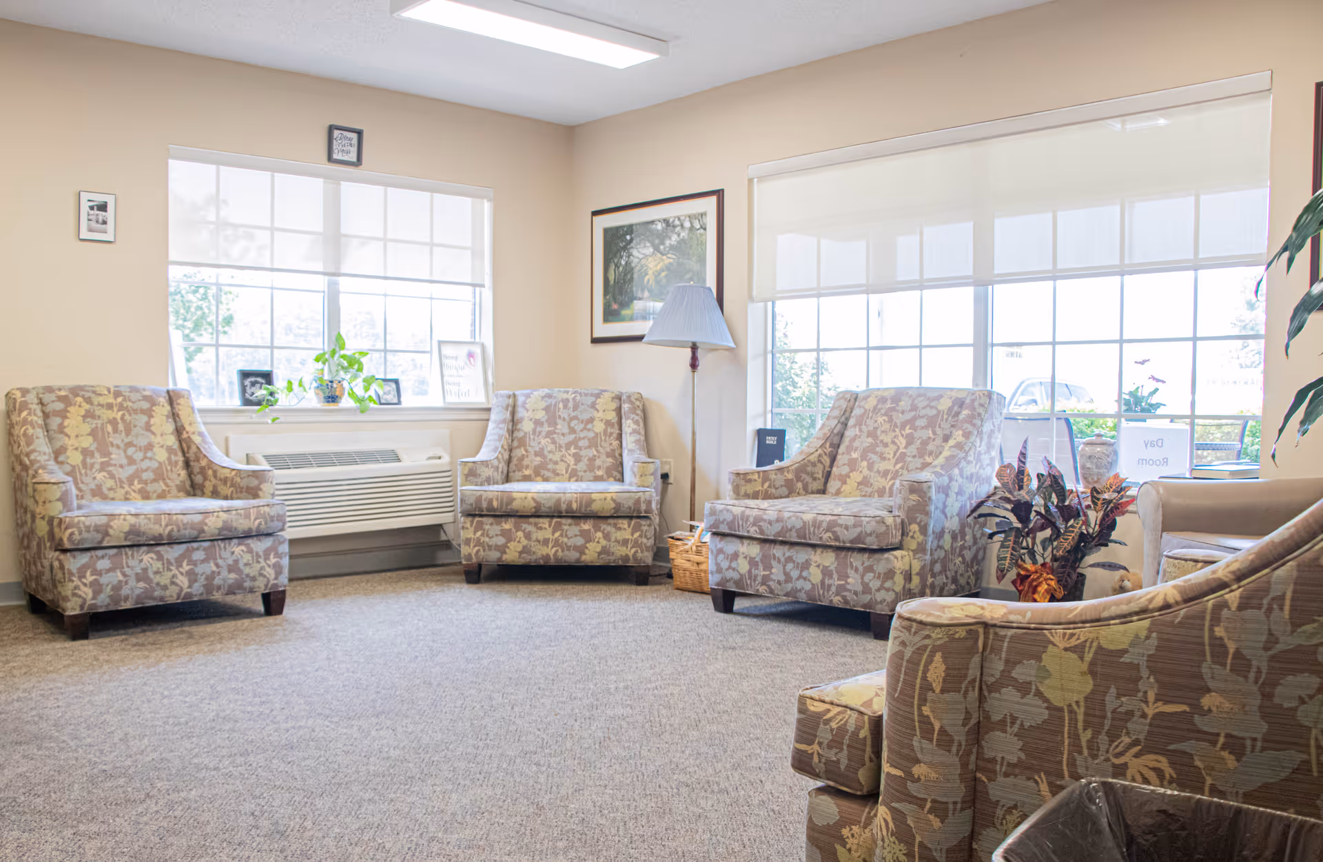 Bright communal sitting room with several patterned armchairs arranged near large windows and a floor lamp.