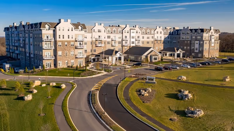 A large multi-story retirement living facility with a combination of stone and light-colored siding exterior. The building has multiple balconies and a covered entrance. The surrounding area includes a curved driveway, parking spaces with cars, and well-maintained green lawns with scattered rocks and small trees under a clear blue sky.