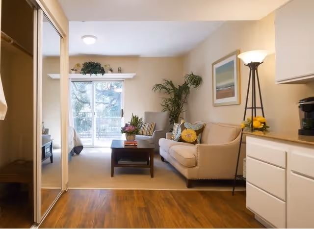 A cozy living room area with a beige sofa adorned with colorful pillows, a gray armchair, a wooden coffee table with a flower arrangement, a tall floor lamp, and a large potted plant. The room has light-colored walls, a framed picture, and a sliding glass door leading to an outdoor balcony. The floor transitions from wood to carpet near the seating area.