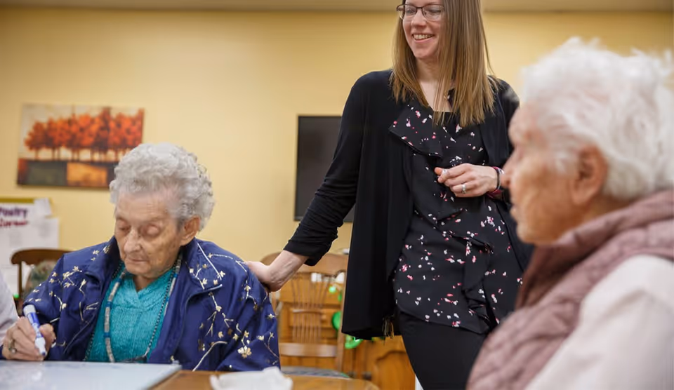 An elderly woman in a blue jacket is writing on a whiteboard at a table, while a younger woman stands beside her smiling. Another elderly woman with white hair and a pink vest is seated nearby, slightly out of focus. The setting appears to be a communal indoor area with warm lighting and a painting of trees on the wall.