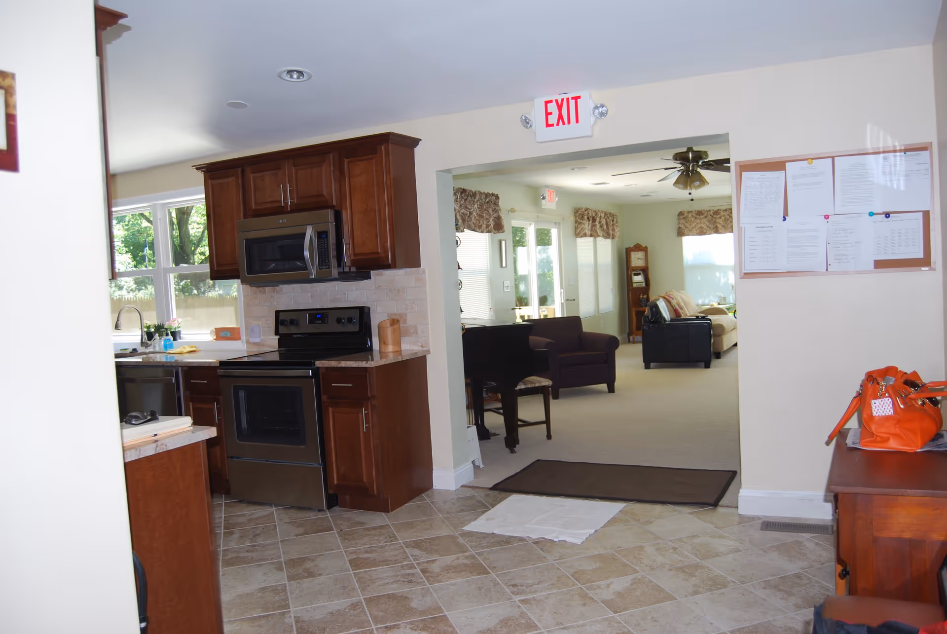 View of a kitchen area with wooden cabinets, a stove, and a microwave. The kitchen opens into a living room with chairs, a piano, and a ceiling fan. There is an exit sign above the doorway and a bulletin board with papers on the right wall. An orange bag is placed on a wooden table near the bulletin board.