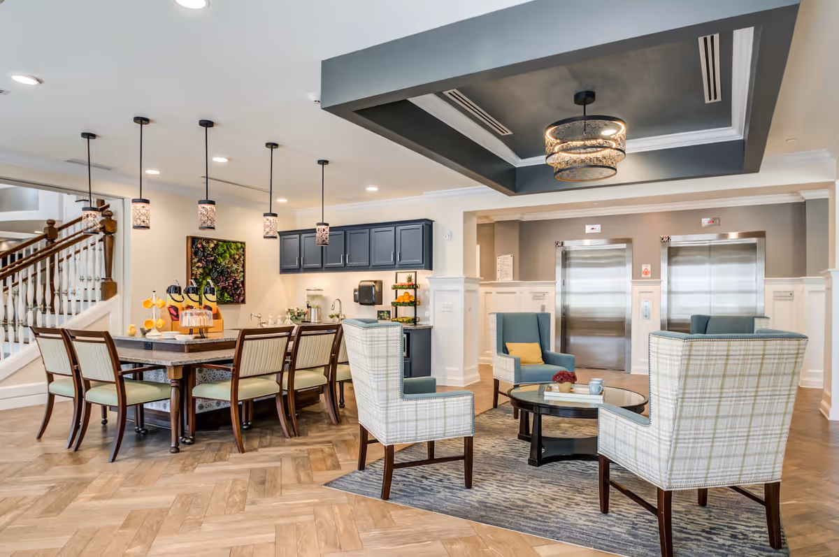A bright and modern common area in a senior living facility featuring a seating area with four upholstered chairs around a round glass coffee table on a rug. Behind the seating area is a large wooden table with six chairs, pendant lights hanging above, and a kitchenette with dark blue cabinets and a countertop with refreshments. Two stainless steel elevators are visible in the background.