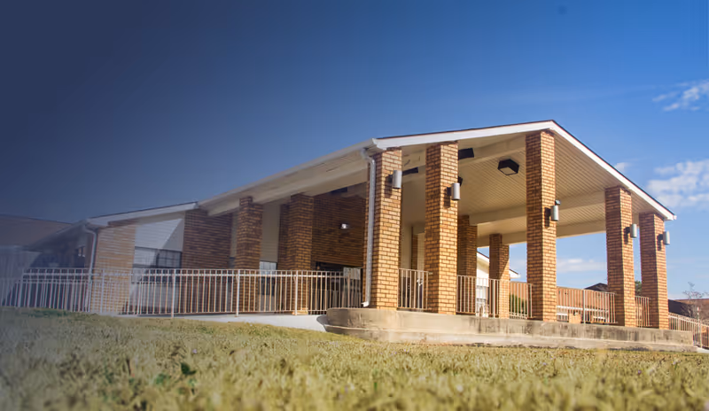Exterior view of a single-story brick building with a covered porch supported by multiple brick columns, surrounded by a metal railing, under a clear blue sky.