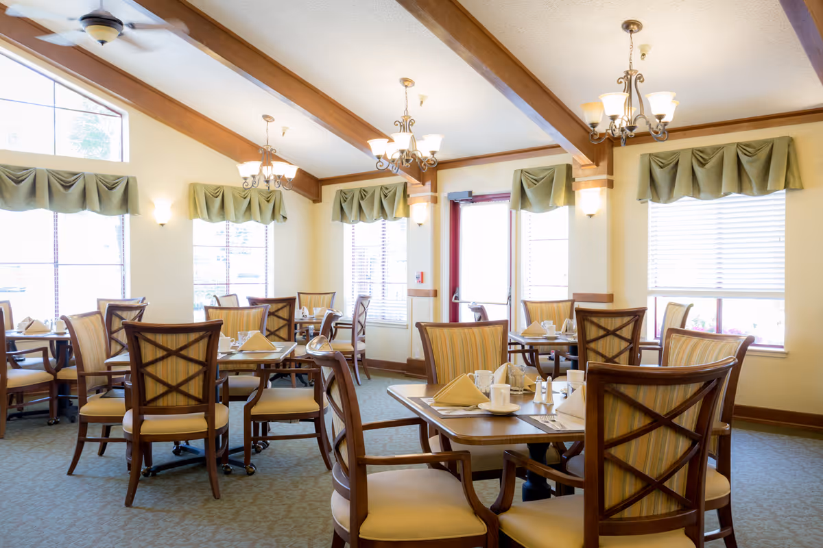 A bright dining room with multiple wooden tables and chairs arranged neatly. Each table is set with folded napkins, cups, and silverware. The room features large windows with green valances, wooden ceiling beams, and elegant chandeliers providing warm lighting.