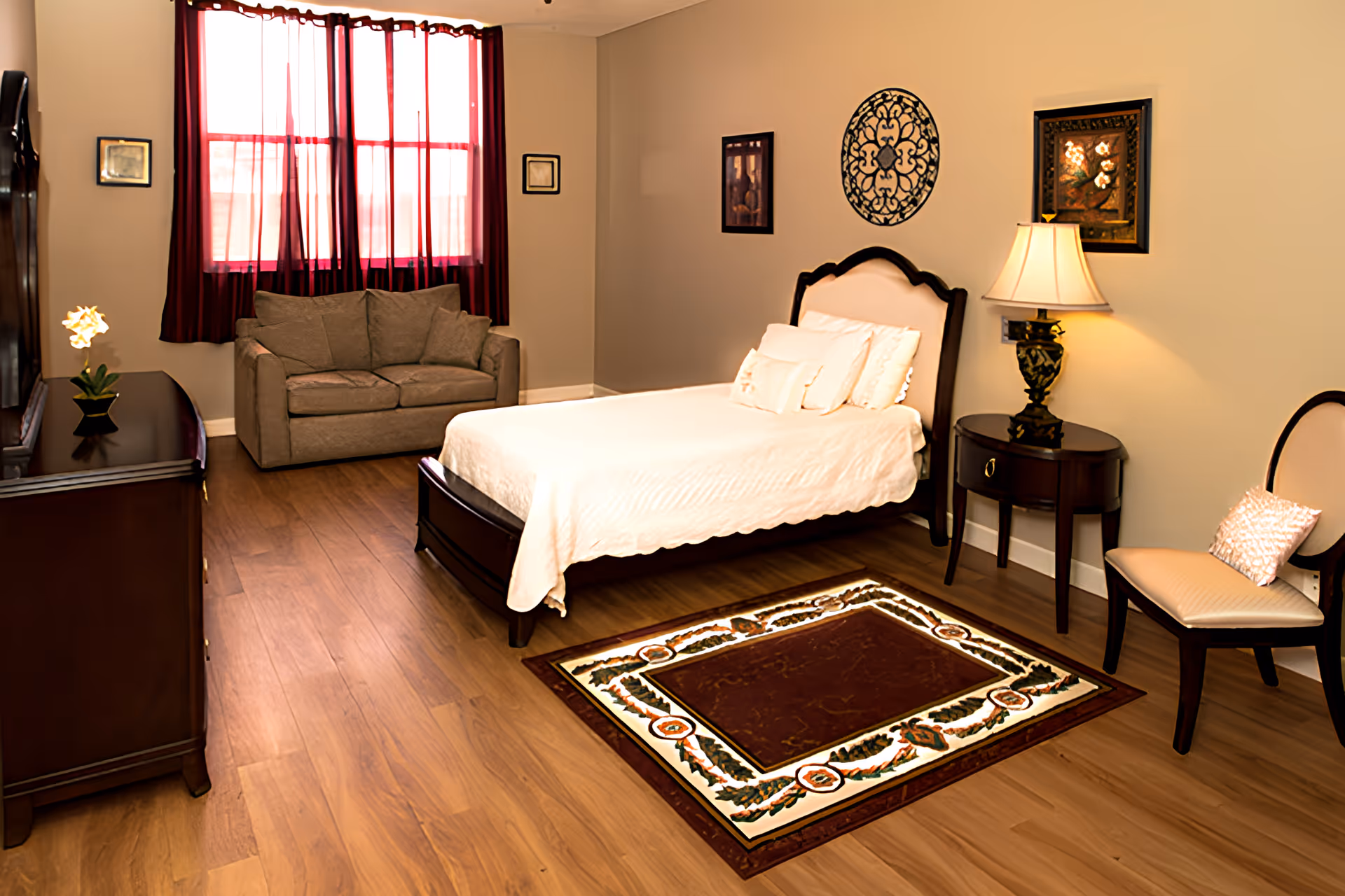 A cozy bedroom with a wooden bed frame and white bedding, a patterned area rug on wooden flooring, a small sofa under a window with red curtains, a wooden dresser with a flower vase, a side table with a lamp, and a chair with a cushion. The walls are decorated with framed artwork and a decorative wall piece.