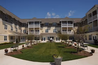 A sunny courtyard area of a senior living facility with a well-maintained lawn, small trees, benches, and potted plants surrounded by a three-story building with balconies and windows.