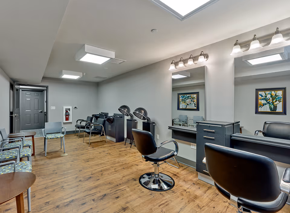 A salon-style interior with styling chairs, mirrors, hair dryers, and a row of waiting chairs in a senior living facility.