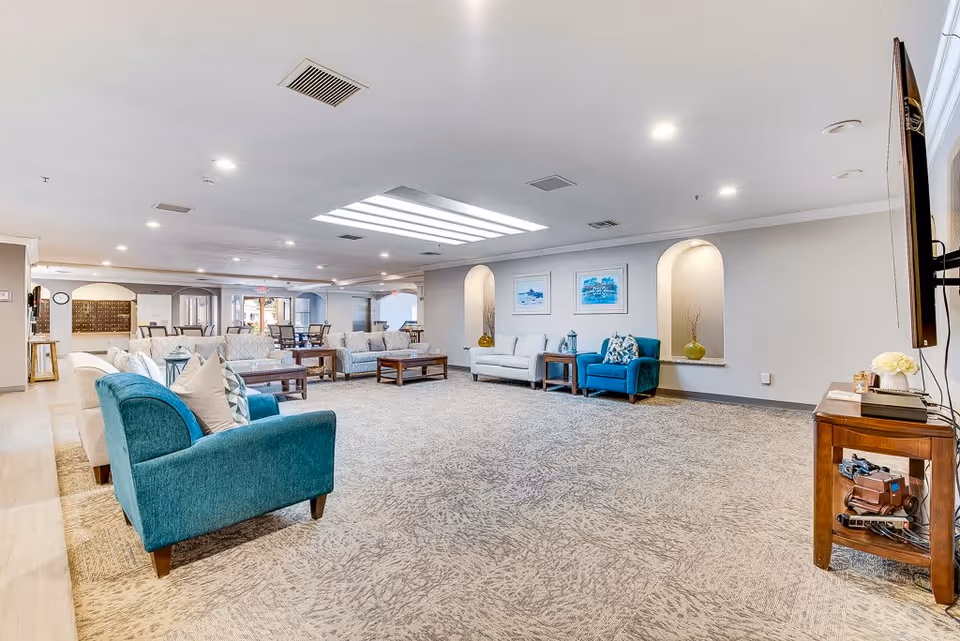 Spacious senior living facility common area with multiple seating options including teal and beige armchairs and sofas arranged around wooden coffee tables. The room has a large carpeted floor, recessed lighting, and a skylight. There are decorative wall niches with vases and framed artwork on the walls. A TV is mounted on the right wall above a wooden side table with various items on it.