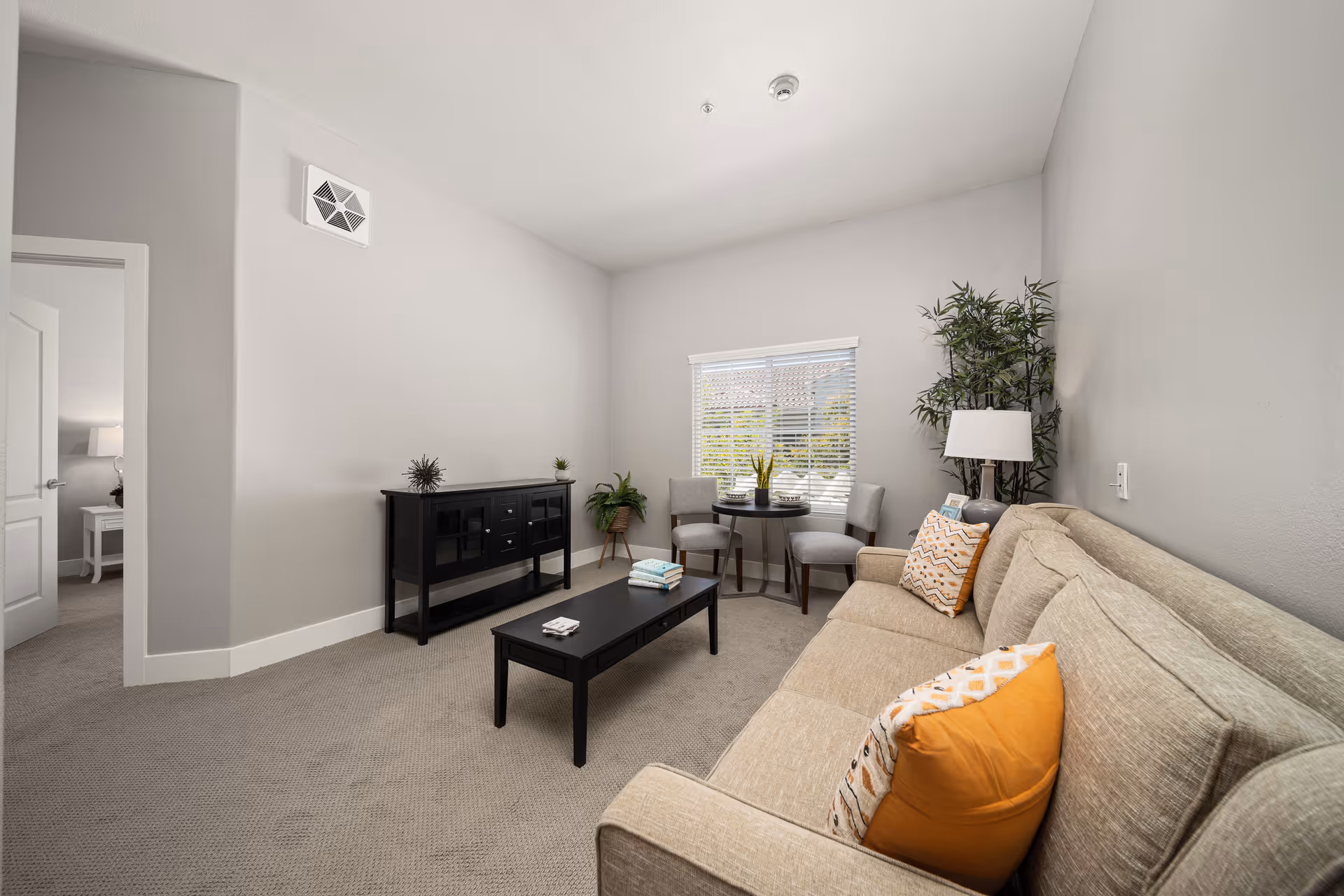 Bright living room with a beige sofa and orange pillows, black coffee table and console, a small round table by the window, and neutral gray walls.