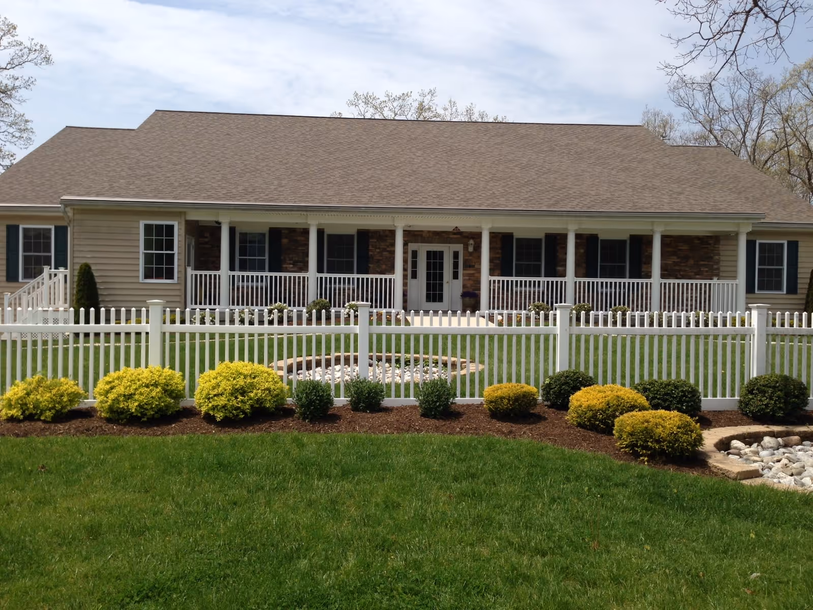 Single-story building with a brown shingled roof, beige siding, and a front porch with white railings. The porch has several columns and a central door with windows on either side. In front of the building is a white picket fence with neatly trimmed bushes and green grass. The sky is partly cloudy.