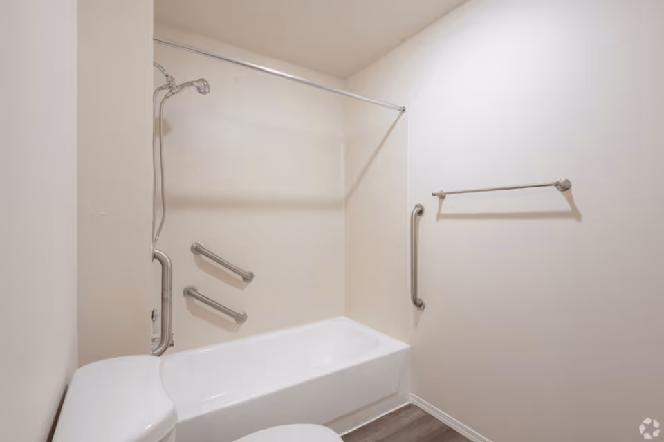 A bathroom with a white bathtub and shower combination, equipped with multiple grab bars for accessibility. There is a showerhead attached to the wall, a towel rack on the adjacent wall, and a toilet partially visible in the foreground. The walls are painted white and the floor has a wood-like finish.