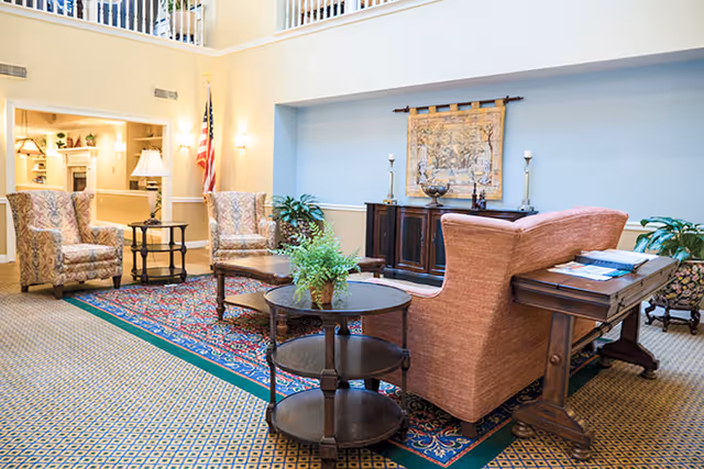 A bright and spacious living room area with patterned carpet and a large decorative rug. The room features a peach-colored sofa, two floral armchairs, a round wooden coffee table with a green plant, and a wooden side table with a lamp. There is a dark wooden cabinet against the wall with decorative items and a tapestry hanging above it. An American flag is visible in the corner near a doorway leading to another room.