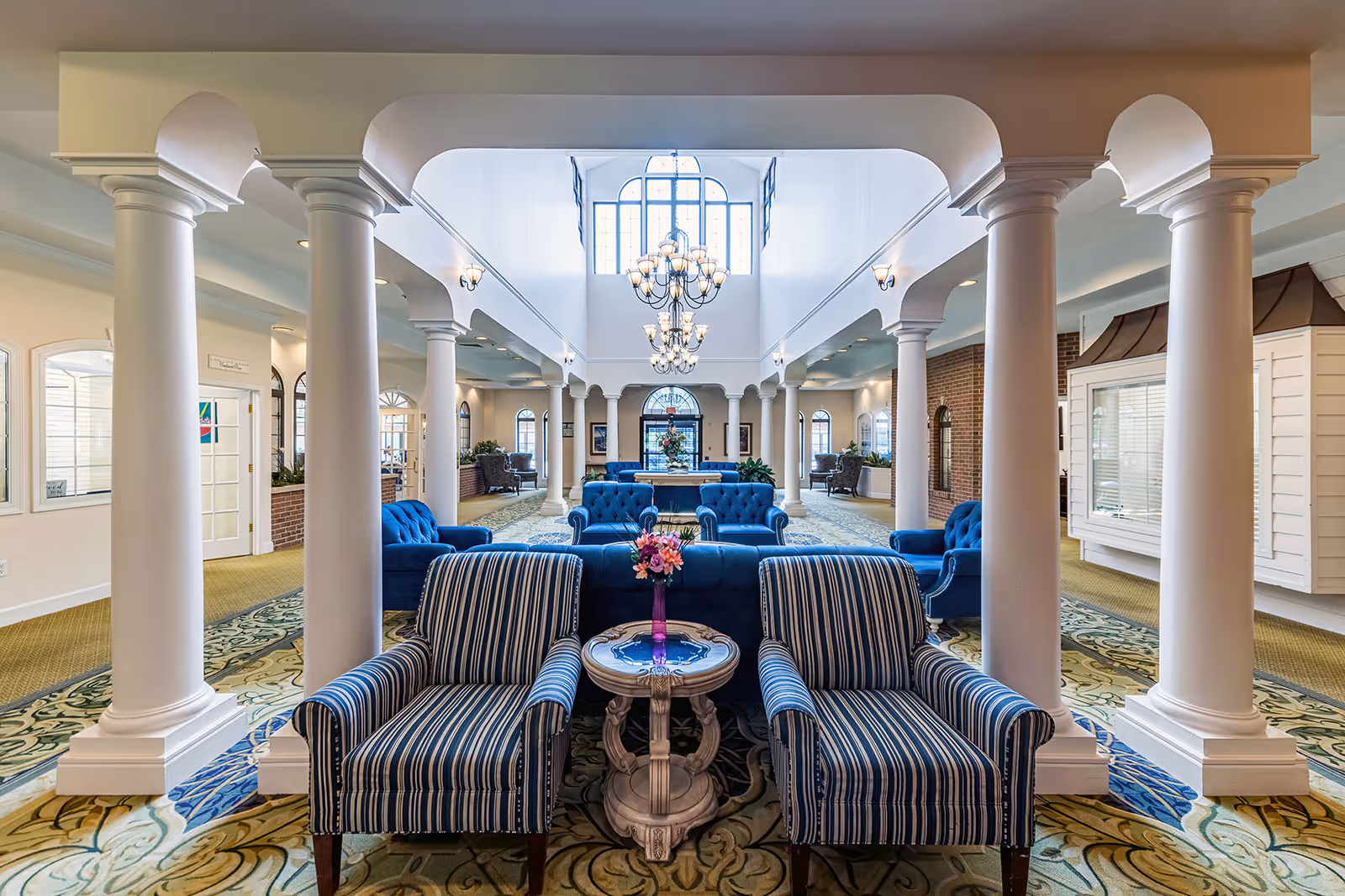 Spacious senior living facility common area with white columns, blue upholstered armchairs and sofas, a decorative carpet, and large windows letting in natural light. A small round table with a vase of flowers is placed between two striped armchairs in the foreground. Elegant chandeliers hang from the high ceiling.