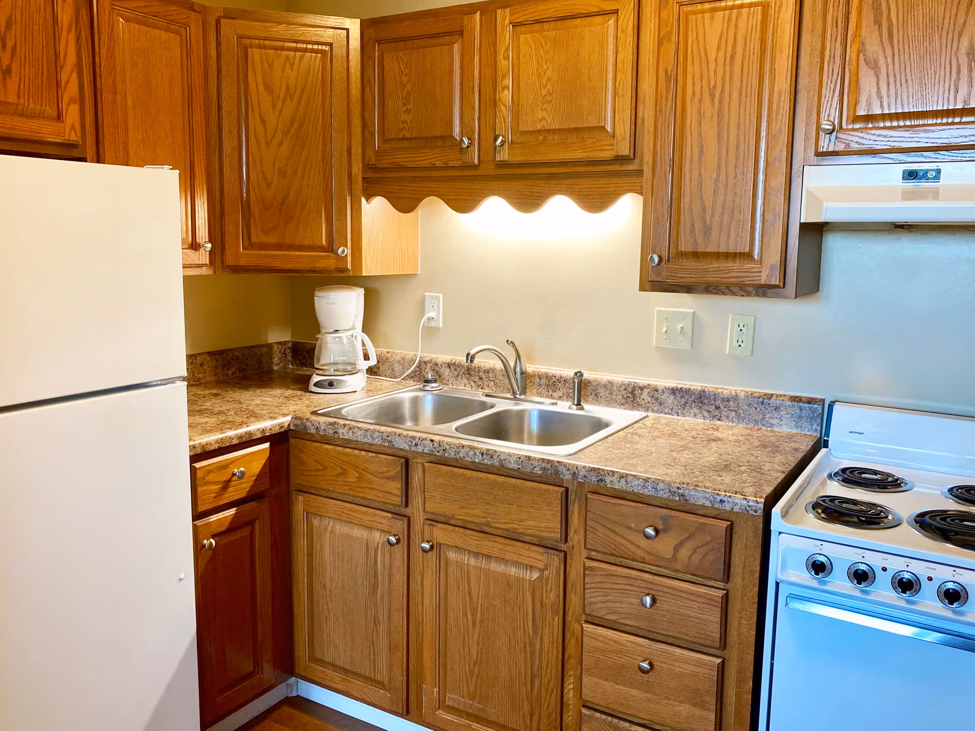 A kitchen area with wooden cabinets, a white refrigerator, a white electric stove with four burners, a double stainless steel sink, and a white coffee maker on the countertop. The countertop has a brown marble pattern and there is under-cabinet lighting illuminating the sink area.