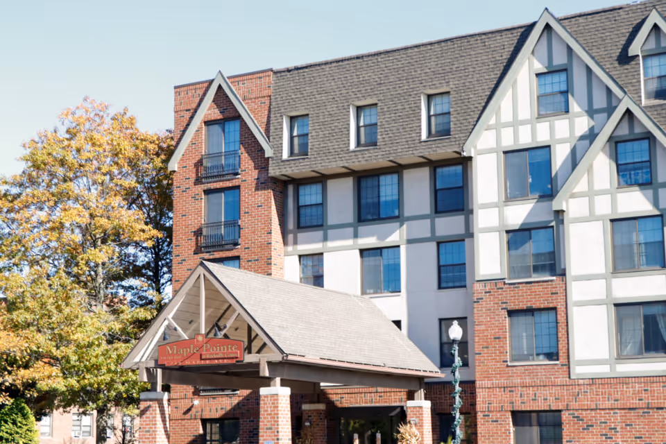 Exterior view of a multi-story senior living facility building with a brick and white panel facade, peaked roofs, and multiple windows. There is a covered entrance with a sign that reads 'Maple Pointe'. Trees with autumn foliage are visible on the left side.